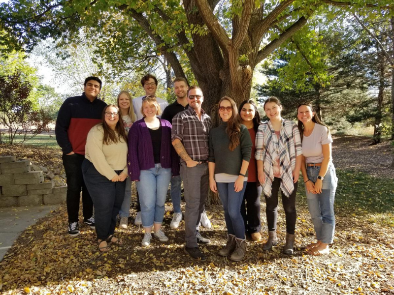 Eleven people pose for a photo in front of a tree